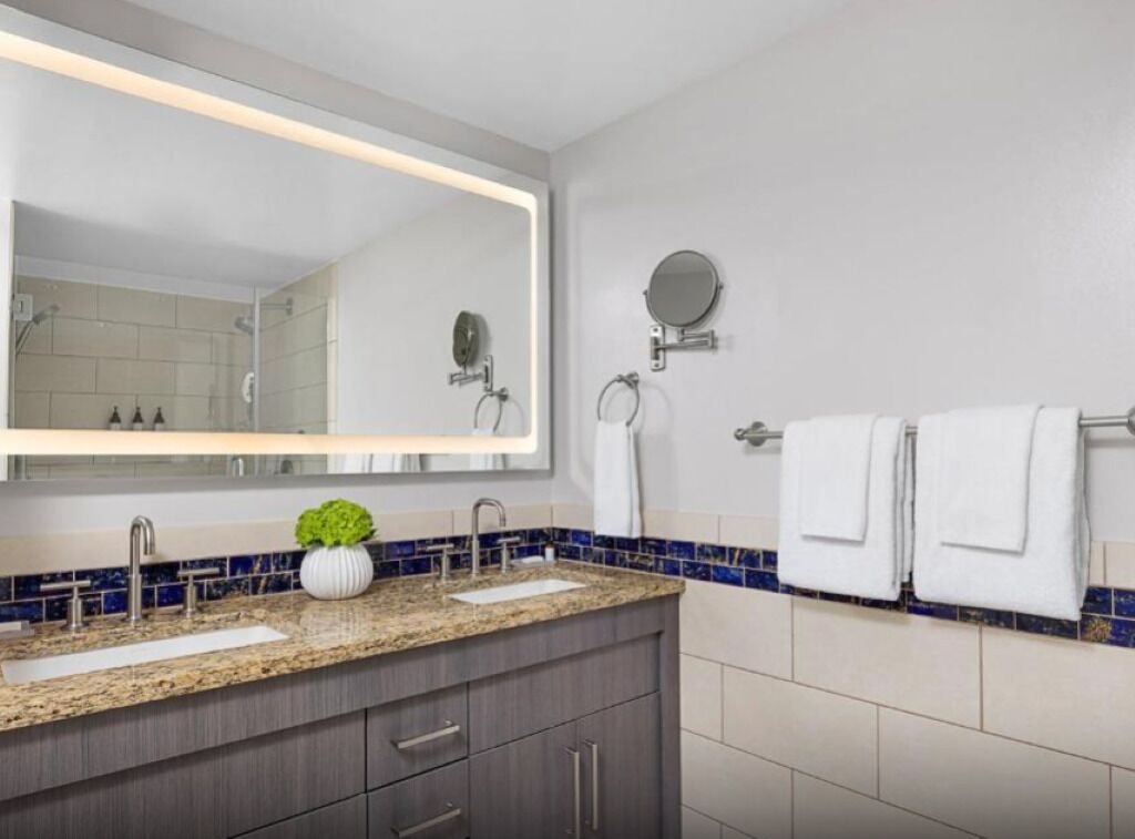 Un baño moderno con un espejo grande, dos lavamanos, un perchero para toallas y una planta pequeña en la encimera.