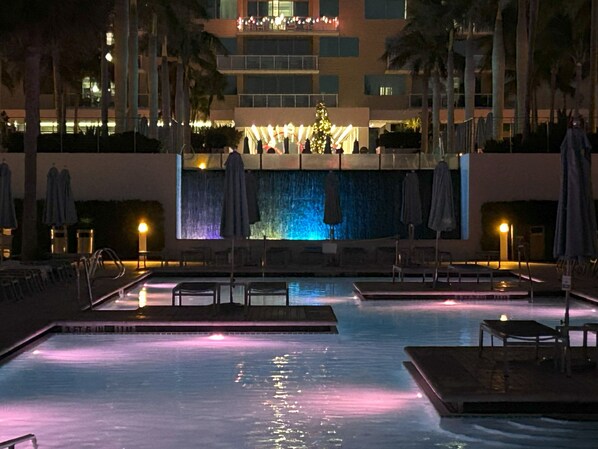 A nighttime swimming pool with a waterfall feature, lounge chairs, and umbrellas.