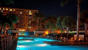 Ein Nachtbild von einem Hotelpoolbereich mit mehreren Schwimmbecken, Palmen und einem Gebäude im Hintergrund, dessen Fenster beleuchtet sind.