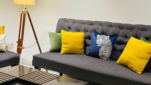 Un canapé gris à motifs, agrémenté de quatre coussins aux couleurs jaune, vert, bleu et à imprimé léopard. Une lampe de sol en bois, avec un abat-jour jaune, se trouve à côté du canapé.