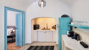 A compact kitchen with a sink, cabinets, and a countertop. There is a door leading to another room, a hanging light fixture, and a shelf with glasses.