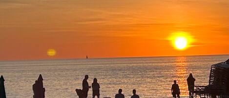 A beach scene with people sitting and standing near the water during sunset.