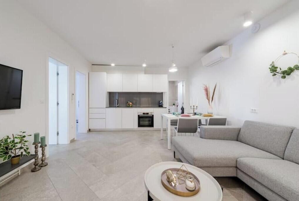 A modern living room with a grey sofa, a coffee table, and a TV on the wall. There is a kitchen area in the background with white cabinets and a dining table.