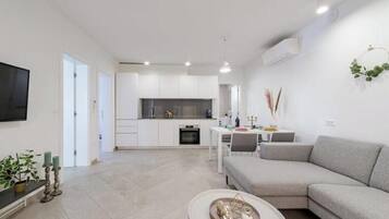 A modern living room with a grey sofa, a coffee table, and a TV on the wall. There is a kitchen area in the background with white cabinets and a dining table.