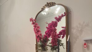 Una consola negra con un espejo, flores rosadas, un florero, una botella y un cuenco decorativo.