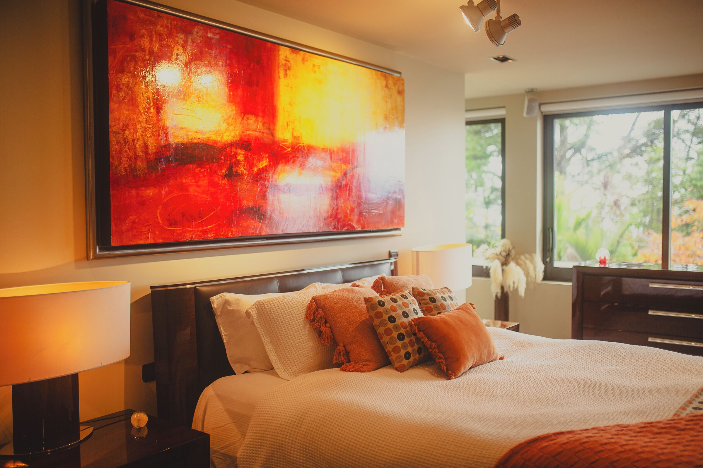 A hotel room with a large abstract painting, a neatly made bed with multiple pillows, a nightstand with a lamp, and a view of trees through a large window.