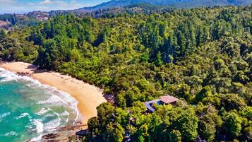 An aerial view of a coastal area with a beach, dense forest, and a small building.