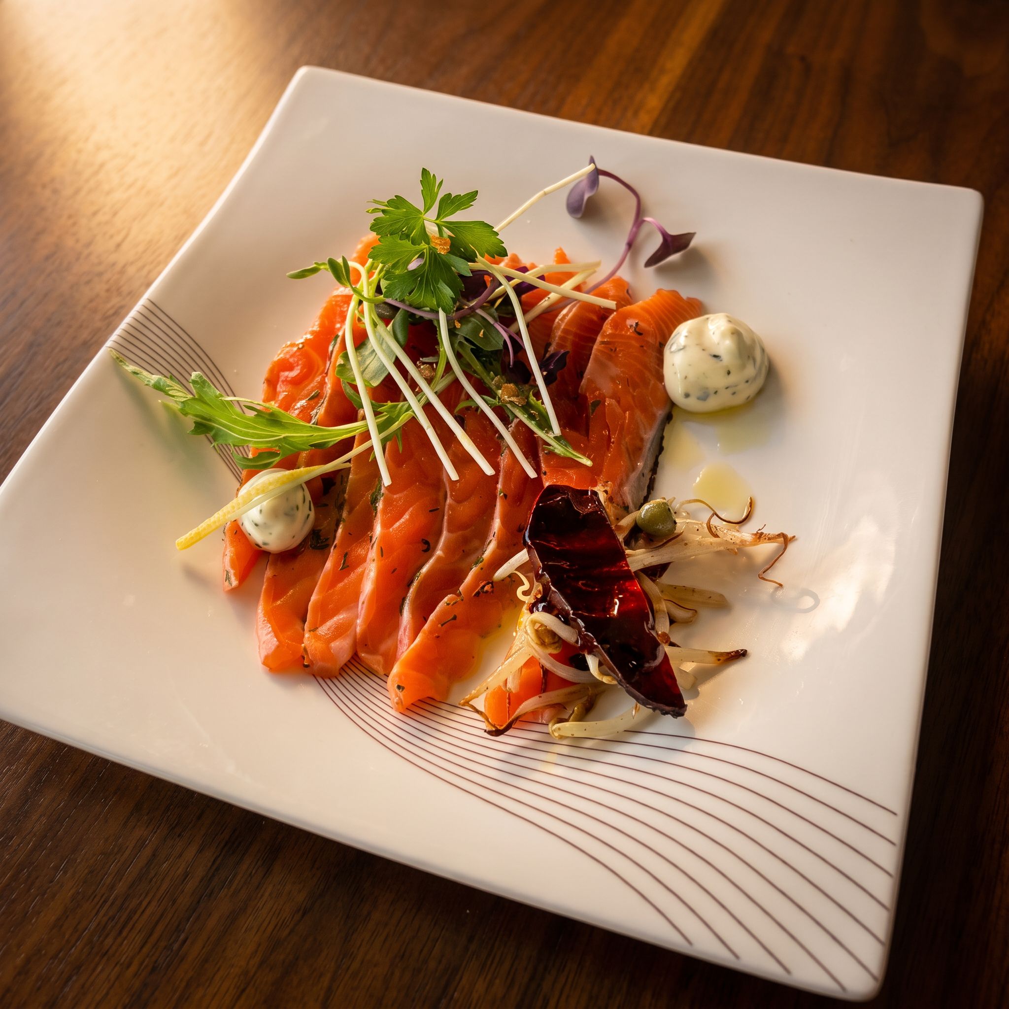 A square plate with salmon slices, sprouts, and a dollop of cream.