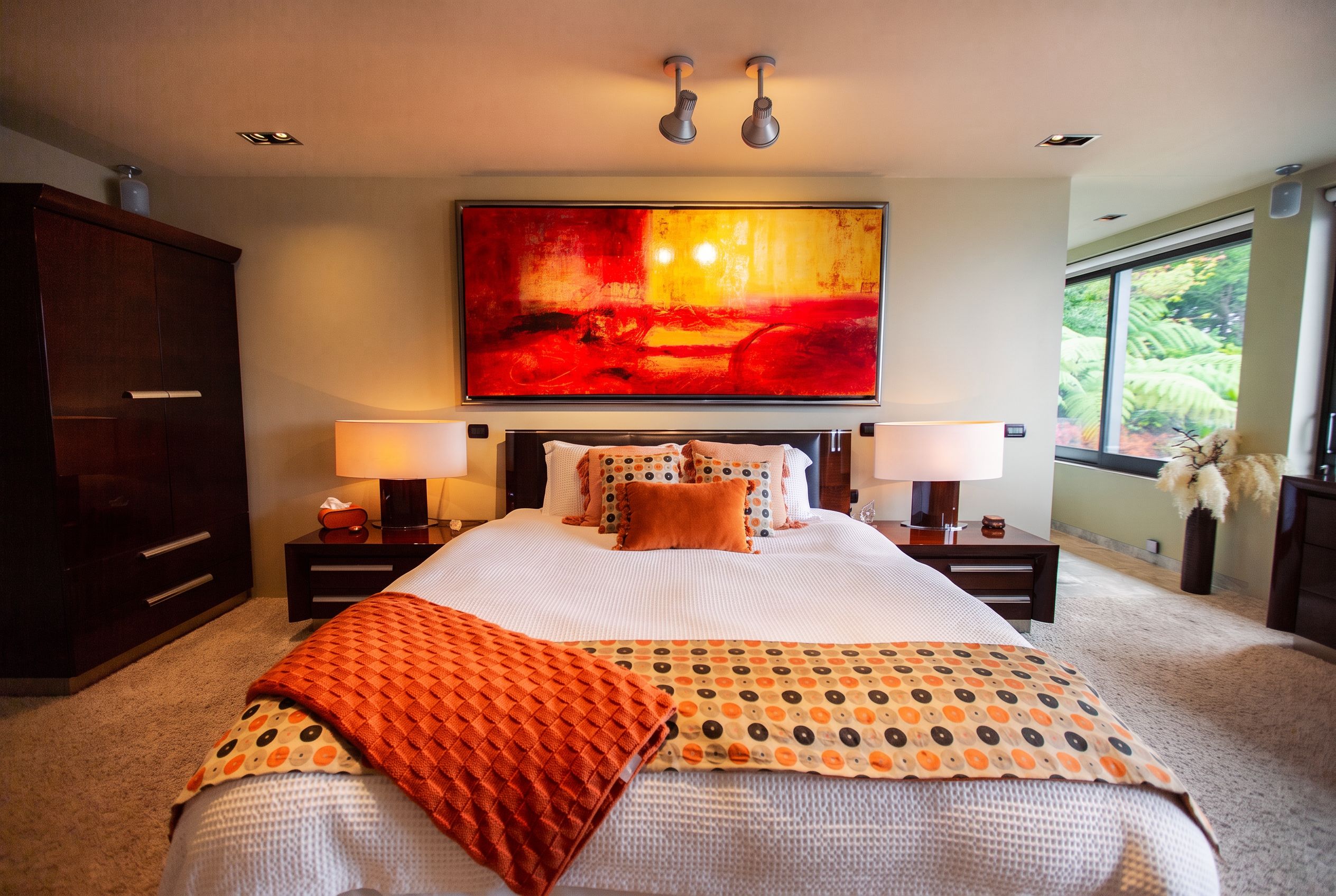A bedroom with a large bed, two bedside tables, a nightstand, a wardrobe, a large abstract painting, and a window with a view of greenery.