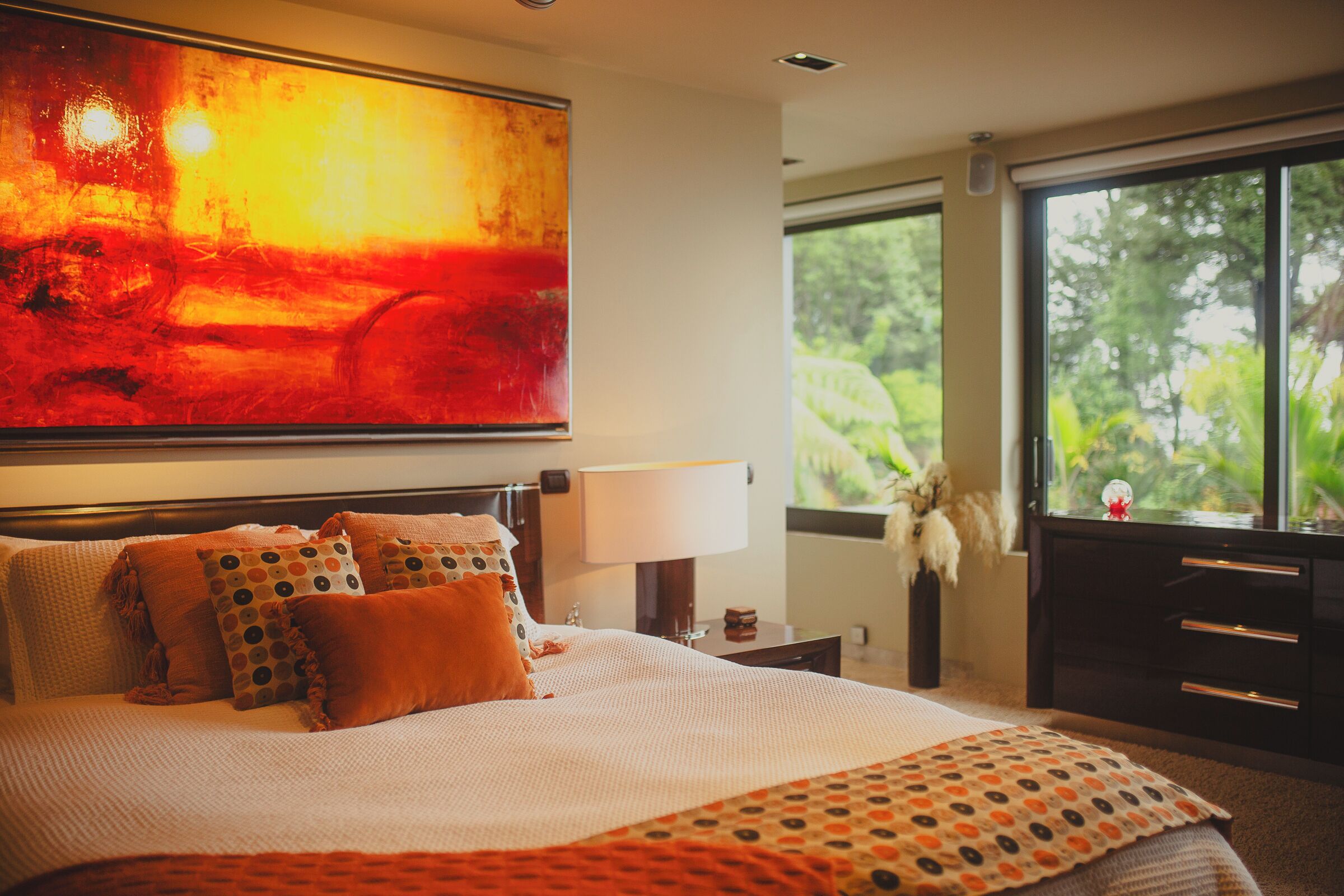 A hotel room with a large bed, a nightstand, a dresser, and a window with a view of greenery.