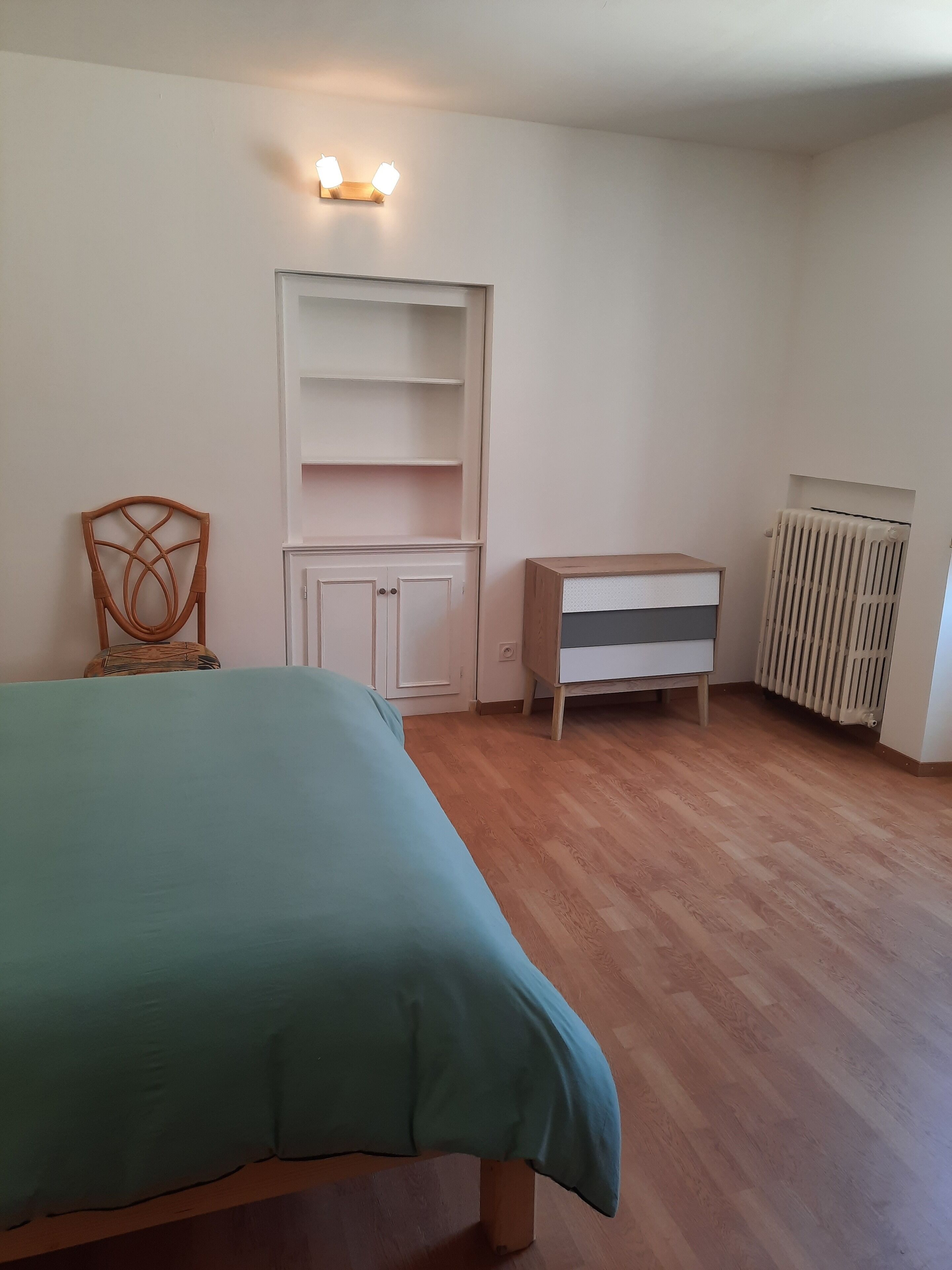 Un dormitorio con cama, una silla, un armario blanco, un radiador y una mesita.