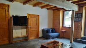 A living room with a television, a sofa, a glass-top coffee table, and wooden walls. - Large apartment in village center with direct access to ski and hiking trails (Prémanon)