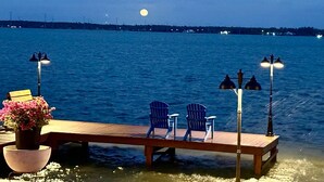 Un muelle de madera con sillas y una mesa, con vista a una masa de agua con una luna en el cielo.