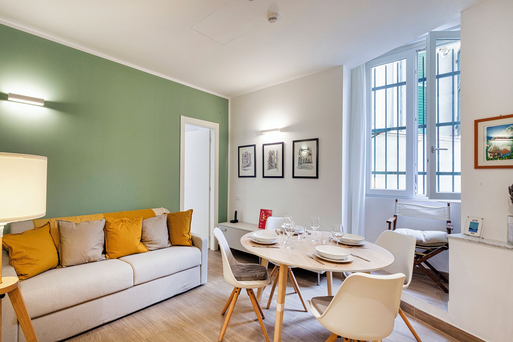 A dining area with a round table set for four, a sofa with cushions, a door, and three framed pictures on the wall.