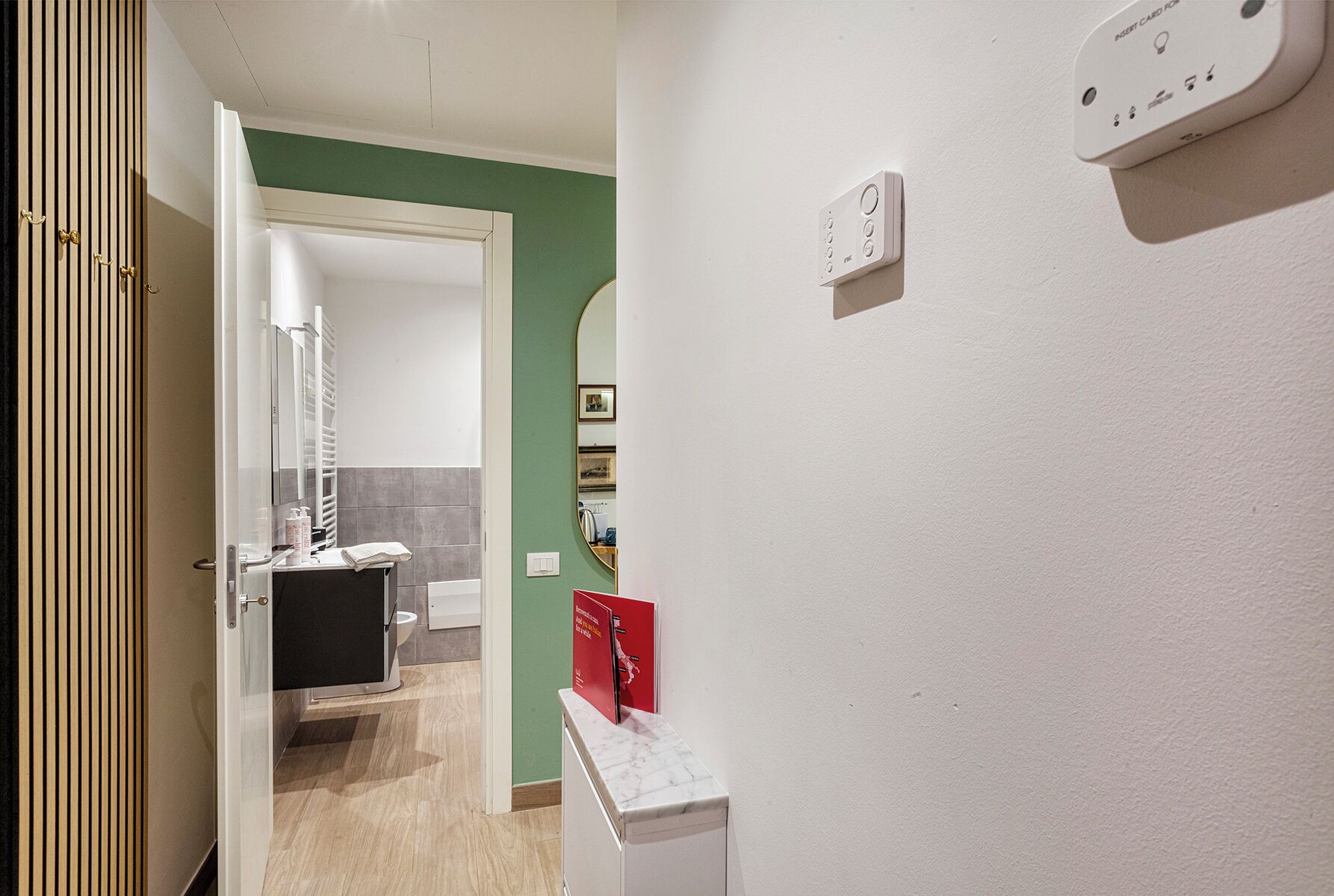 Une chambre d’hôtel avec une porte en bois, un mur blanc muni d’un thermostat, un miroir et un petit panneau rouge.