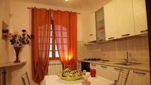 A kitchen with white cabinets, a stove, and a sink. There is a window with orange curtains, a vase with flowers, and a table with a yellow bowl and a red cup. - Cozy mini-apartment ideal for a couple. (Tropea)