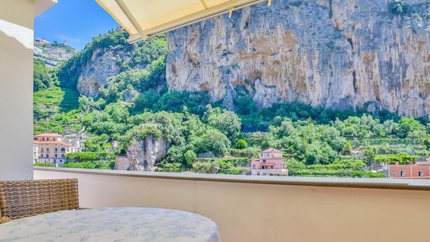 A balcony with a view of a cliff and greenery, a wicker chair, and a table with a floral tablecloth.
