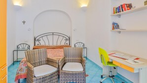 A room with a bed, two wicker chairs, a desk with a chair, and bookshelves.