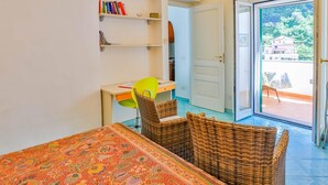 A room with a bed, a desk, a chair, and a bookshelf. There is a door leading to a balcony with a view of buildings and trees.