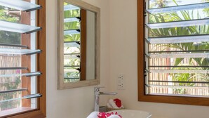 Un baño con lavamanos blanco, un espejo y dos ventanas con persianas.