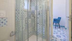 A bathroom with a glass-enclosed shower, patterned tiles, a toilet, and a blue chair in the adjacent room.