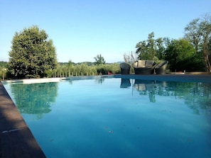 A clear swimming pool with a wooden deck edge, surrounded by trees and a modern outdoor seating area.