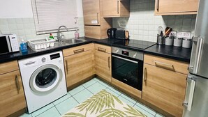 A compact kitchen with wooden cabinets, a built-in washing machine, a microwave, a toaster, and a refrigerator. - 3bedroom- Near to Station-private Garden-wifi (Liverpool)