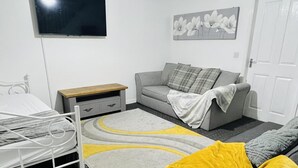 A bedroom with a bed, a grey sofa, a wooden nightstand, a TV mounted on the wall, and a door. - 3bedroom- Near to Station-private Garden-wifi (Liverpool)