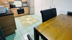 A compact kitchen with wooden cabinets, a refrigerator, and a dining area with a table and chairs. - 3bedroom- Near to Station-private Garden-wifi (Liverpool)