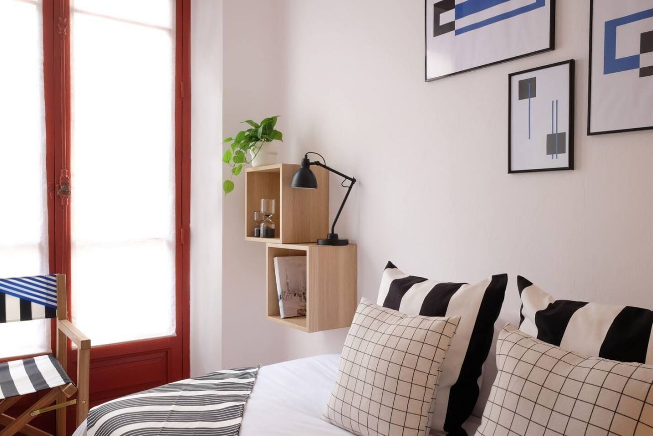A bedroom with a bed, pillows, a desk with a chair, a hanging shelf with a plant, and framed artwork on the wall.
