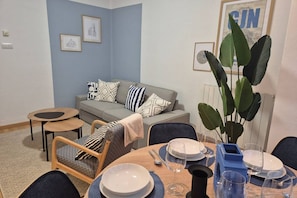 A living room with a grey sofa, a dining table set with plates and glasses, a coffee table, and a large plant.