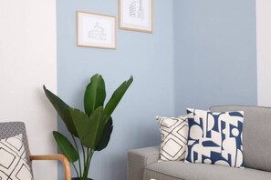 A room with a grey sofa, patterned cushions, a potted plant, and framed pictures on the wall.