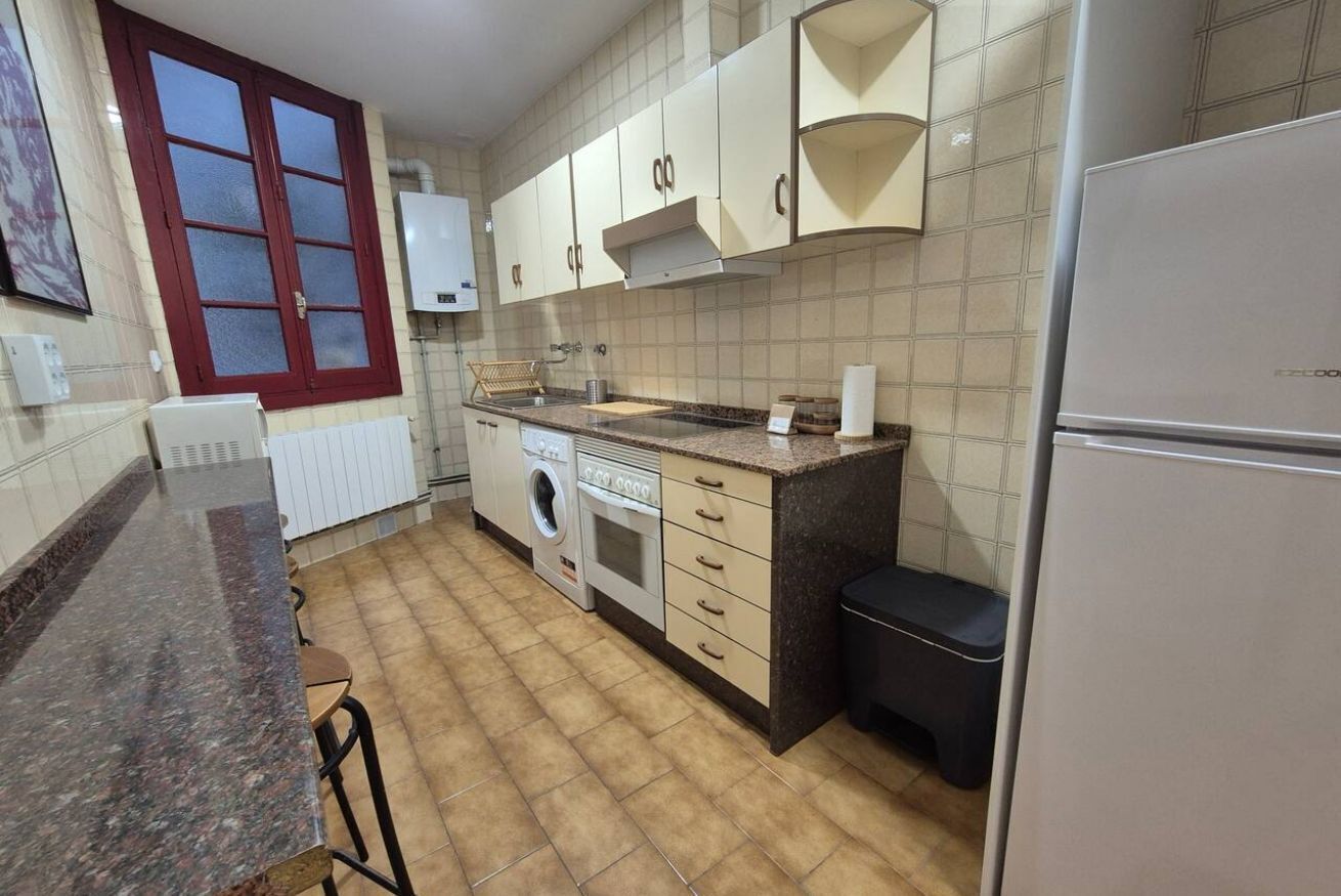 A kitchen with a washing machine, a refrigerator, and a window with a red frame.