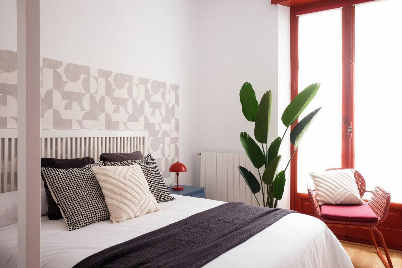 Une chambre à coucher avec un lit, des oreillers, une lampe rouge, une plante et une chaise près d’une fenêtre.