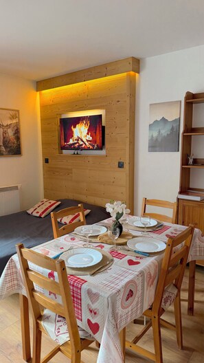 Un coin repas avec une table dressée pour quatre personnes, un téléviseur fixé au mur au-dessus de la cheminée et une bibliothèque sur le côté droit.