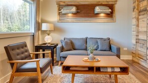Een gezellige woonkamer met een bank, fauteuil en salontafel. Er is een raam met uitzicht op bomen, en een houten plank met twee lampen.