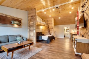 A cozy cabin interior with a loft bed, a sofa, a wooden coffee table, and a kitchen area. - Suite 3 – The Harris Inn in Harrison Hills (Tomahawk)