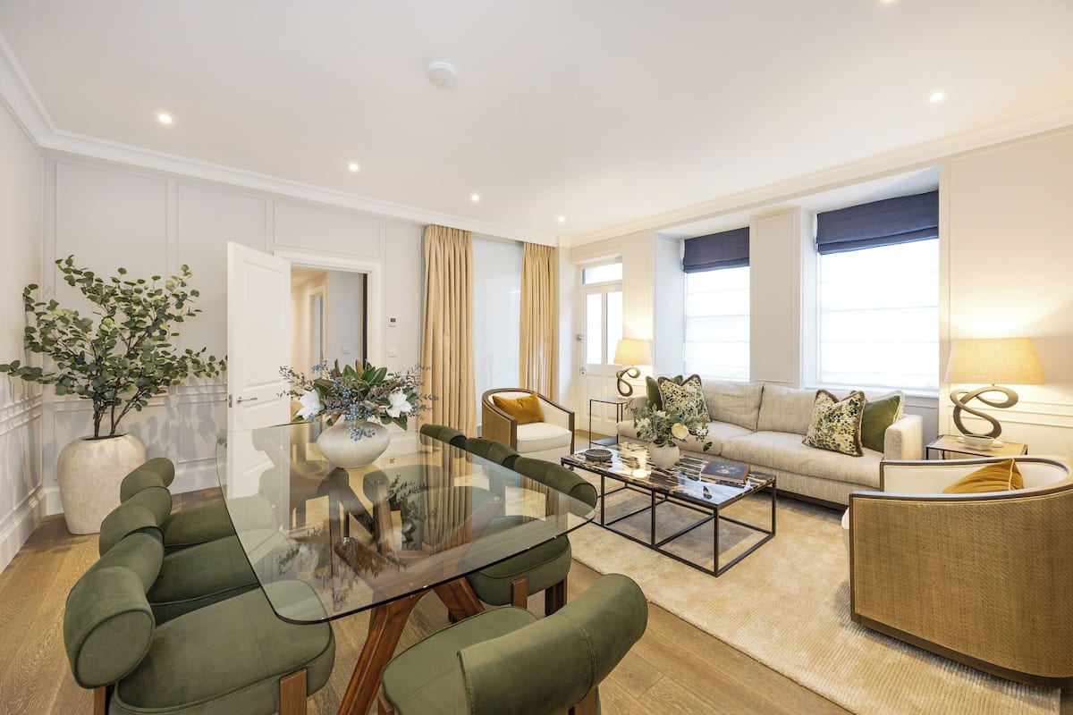 A modern living room with a glass-top dining table, green upholstered chairs, a beige sofa with a patterned throw, and a side table with a lamp.