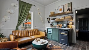 Un salon chaleureux avec un canapé en cuir marron, une table basse ronde et un tapis en peau de vache. On aperçoit également un coin cuisine avec des meubles de couleur turquoise et un amplificateur Marshall.