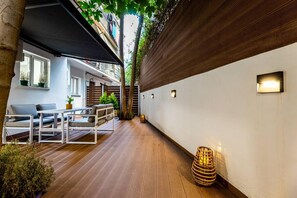 A narrow outdoor corridor with wooden flooring, a white wall on one side, and a black awning on the other. There are small wall-mounted lights and a wicker basket on the floor.