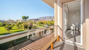 Un balcón con piso de madera y baranda de vidrio que da a un paisaje verde y edificios a lo lejos.