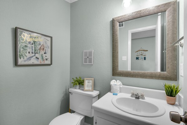 Un baño con inodoro, lavabo y espejo. Hay un cuadro enmarcado en la pared y una pequeña planta sobre el lavabo.
