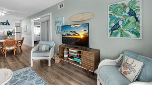 Een woonkamer met een flatscreen televisie, een houten tv-meubel en een eethoek met rieten meubilair.