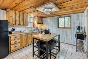 A rustic kitchen with wooden cabinets, a black refrigerator, a wooden dining table with two chairs, and a hanging light fixture.