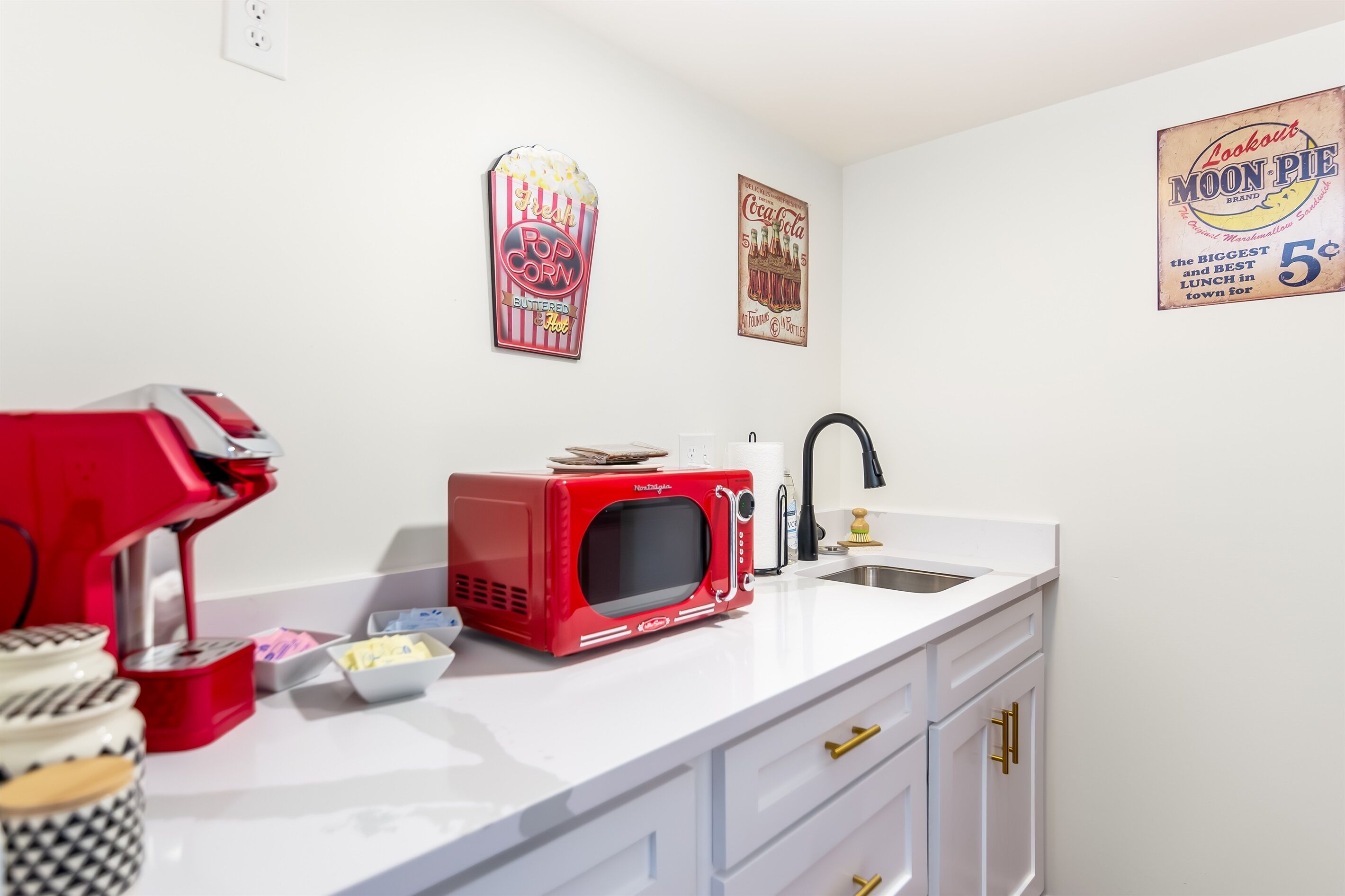 Une cuisine avec un micro-ondes rouge, une machine à café et un évier. On y trouve des décorations murales et un pot de beurre posé sur le comptoir.