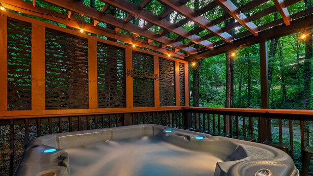 A hot tub under a wooden pergola with string lights, surrounded by a forested area.