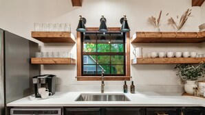 A kitchen with a stainless steel sink, wooden shelves, and a window with a view of greenery.