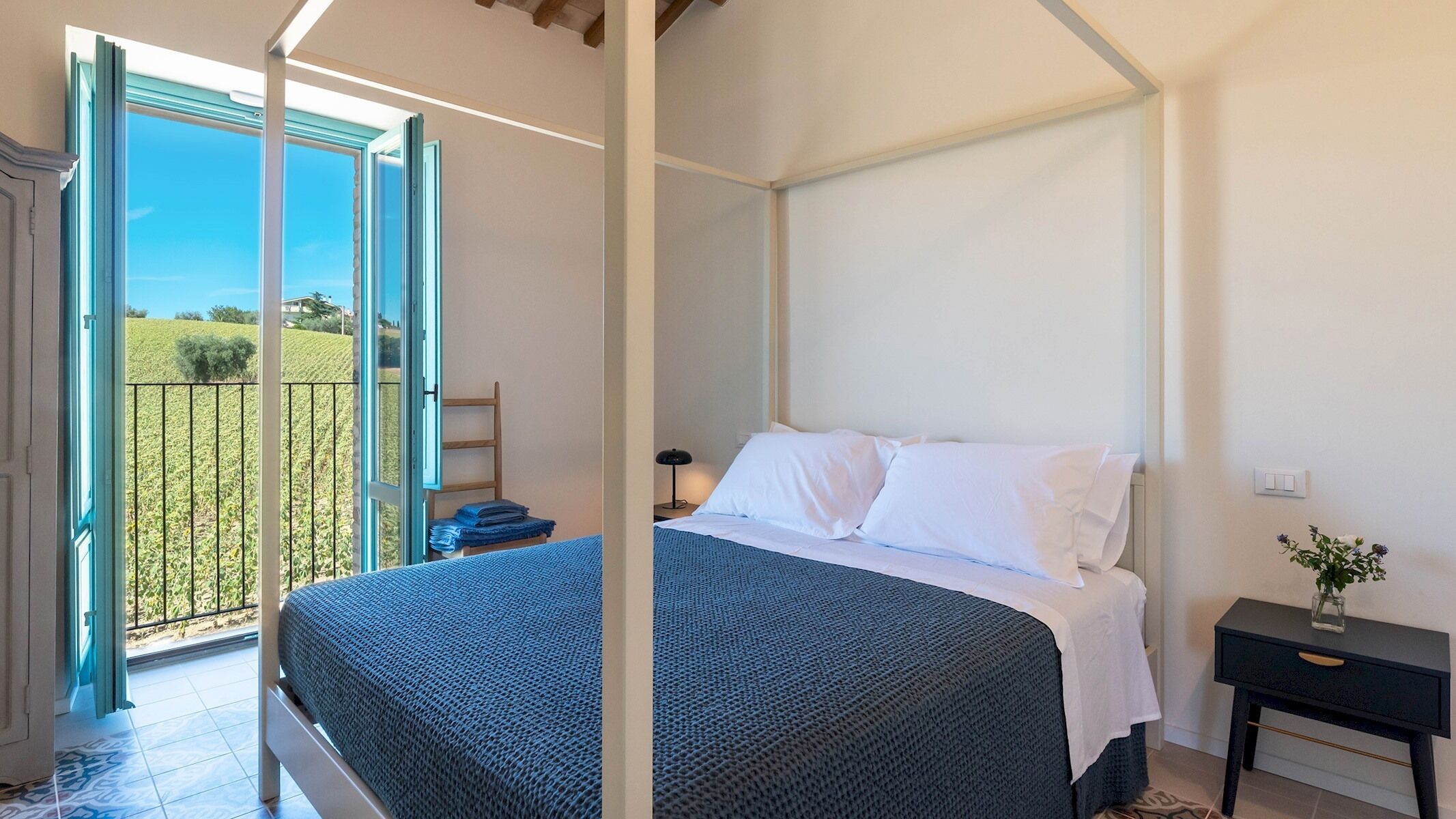 A bedroom with a four-poster bed, a nightstand, a lamp, and a view of a green landscape through an open door.