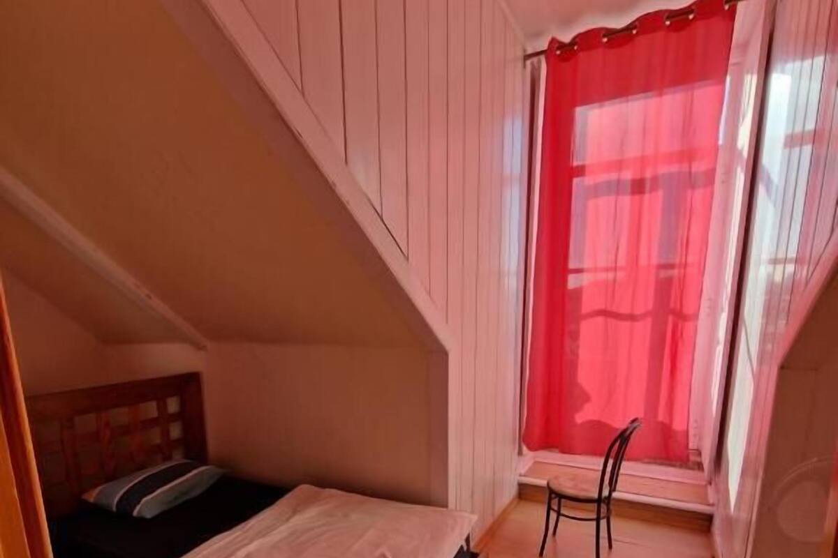 A small, cozy bedroom with a single bed, a chair, and a window with red curtains.