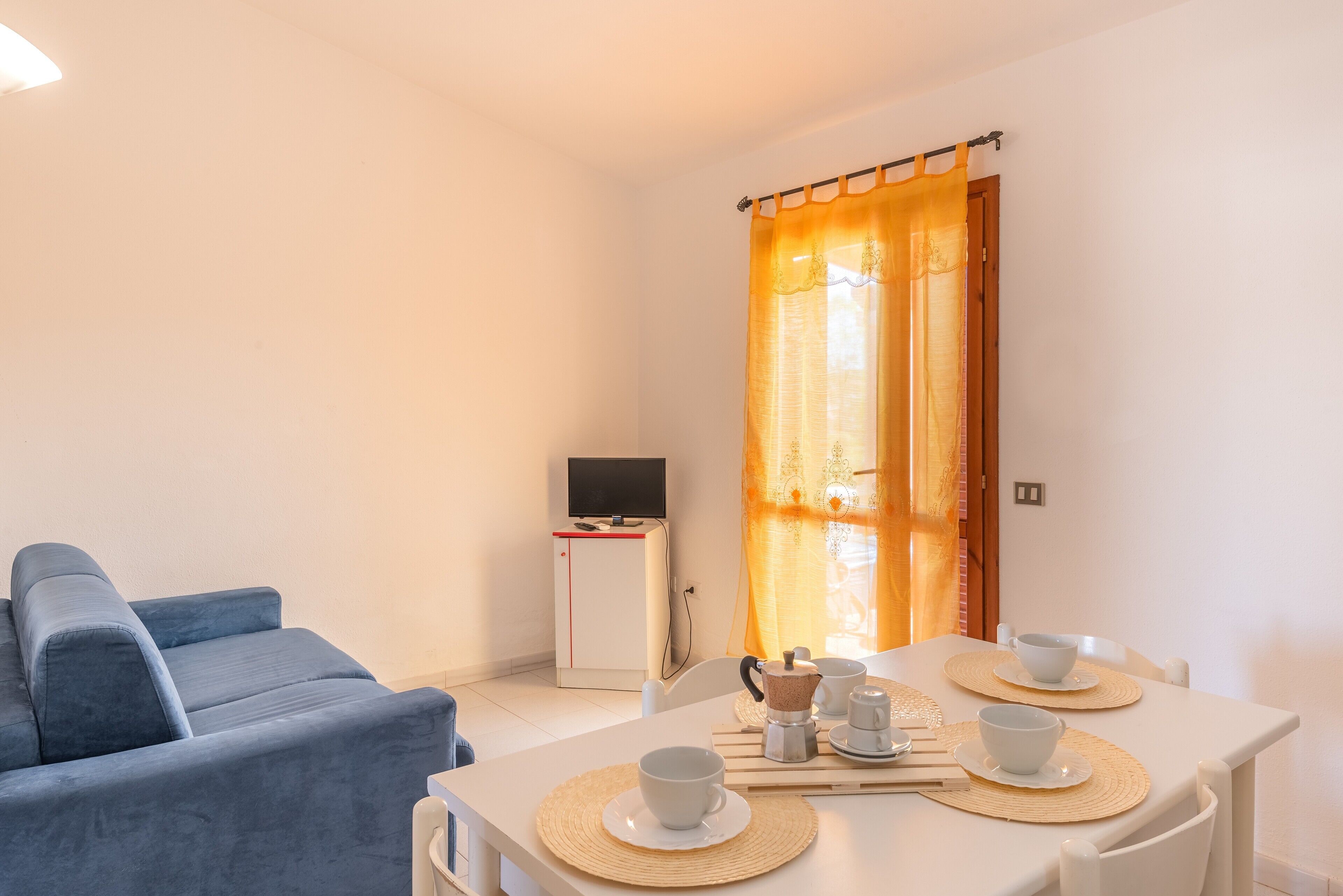 Una sala moderna con un sofá, una mesa de comedor con tazas y una cafetera, y un televisor sobre un mueble bajo.